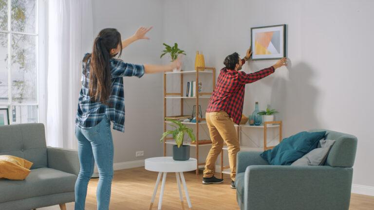Happy Couple Hanging Picture on the Wall, Boyfriend Moves It, Girlfriend Tells Him when the Frame is Hanging Straight. Funny Moment in Young Couple's Life. Modern Stylish Apartment