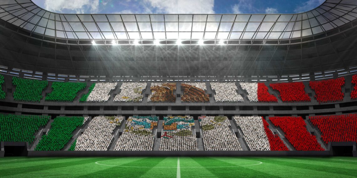 Vista interior de un estadio de fútbol lleno de aficionados formando la bandera de México en las gradas, bajo un cielo parcialmente nublado.