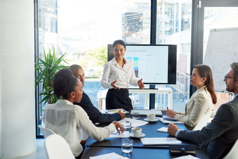 Grupo de profesionales en junta laboral discutiendo estrategias sobre el regreso a la oficina, con presentación en pantalla y liderazgo femenino al frente