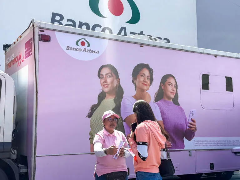 Representante de Banco Azteca conversa con una mujer frente a una unidad móvil rosa de la campaña SOMOS Prevención.